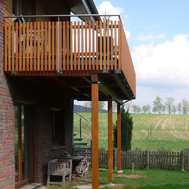 Holzbalkon mit Geländer, erstellt von einer Bautischlerei, stützt sich auf robuste Säulen, mit ländlicher Aussicht.