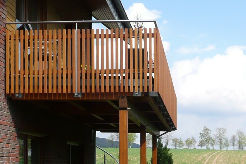 Holzbalkon mit Geländer, erstellt von einer Bautischlerei, stützt sich auf robuste Säulen, mit ländlicher Aussicht.