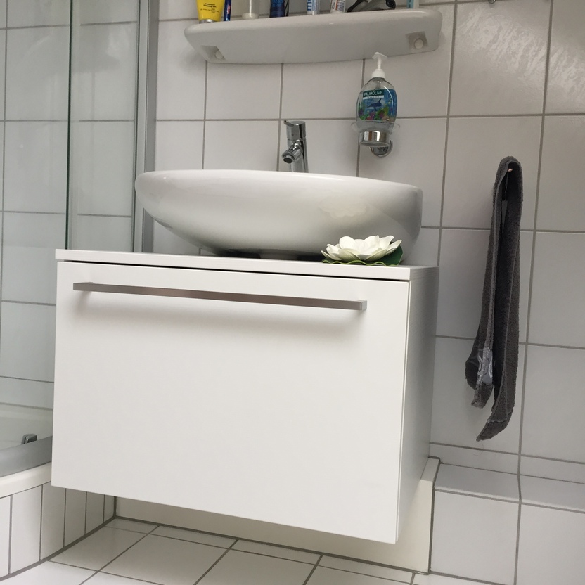 Ein minimalistisches Badezimmer mit einem weißen Waschbecken auf einem maßgeschneiderten Badmöbel nach Maß. Helle, saubere Fliesen runden das Design ab.