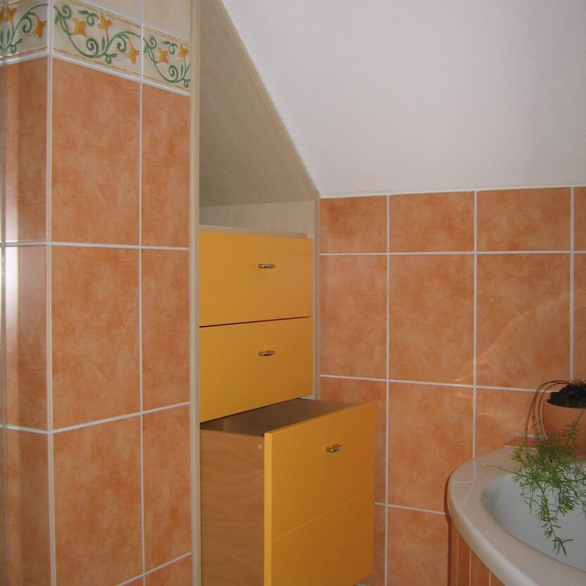 Ein gelber Eck-Badezimmerschrank, optimal als Badmöbel nach Maß unter einer Dachschräge positioniert, umgeben von terrakottafarbenen Fliesen.