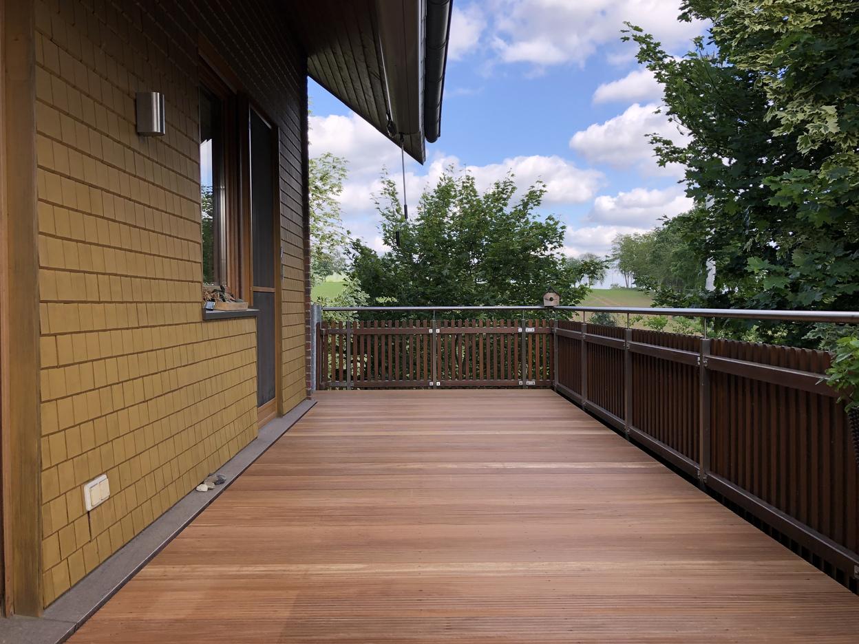 Weitläufige Holzterrasse mit Geländer, Blick auf Bäume und blauen Himmel.