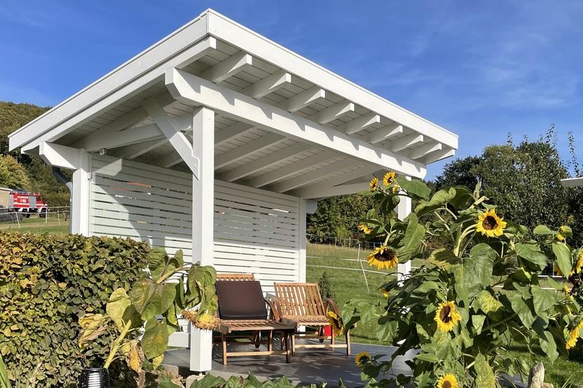 Moderne wei&szlig;e Gartenlaube mit Holzm&ouml;beln, umgeben von bl&uuml;henden Sonnenblumen und Gr&uuml;nfl&auml;che.